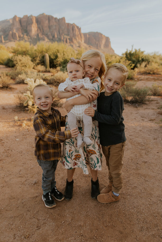 Three adorable oldest children hug their baby sister while taking family photos in the desert of Arizona. #Arizonfamilyphotographer #Phoenixphotolocations #superstitionmountains #familyphotographer #ChelseyMichellePhotography #Scottsdalephotographer #familyphotosinspo #LostDutchmanStatePark