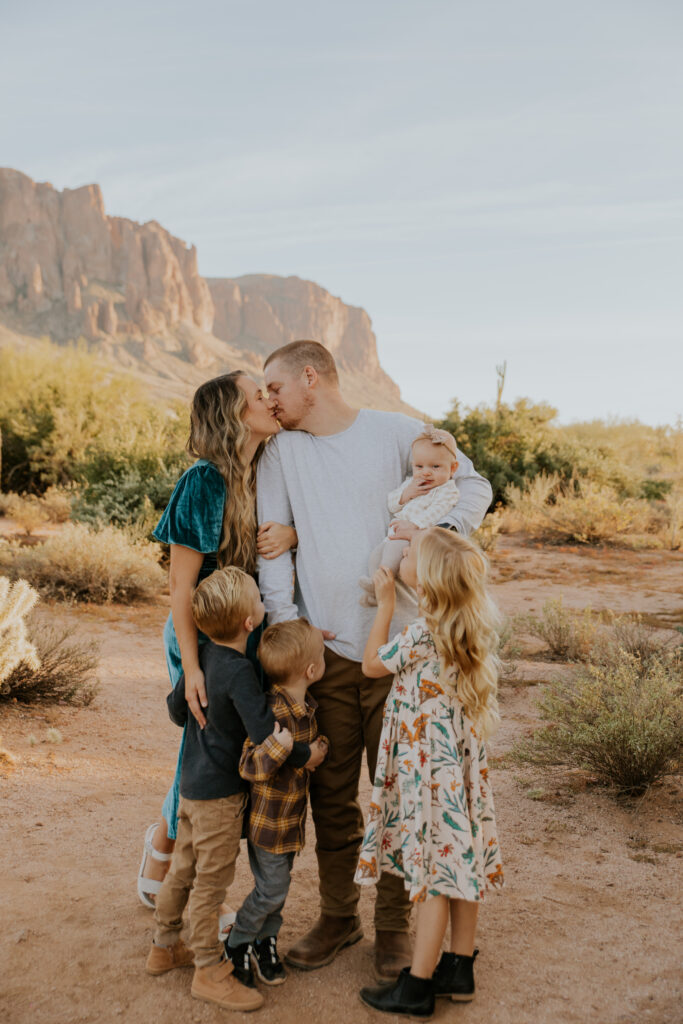 A cute moment where mom and dad are kissing and their young kids watch while giggling during their Arizona family pictures. #Arizonfamilyphotographer #Phoenixphotolocations #superstitionmountains #familyphotographer #ChelseyMichellePhotography #Scottsdalephotographer #familyphotosinspo #LostDutchmanStatePark