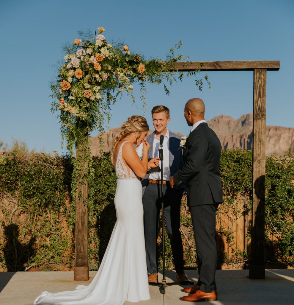 The bride and groom stand with the officiant in front of a floral arrangement with the Superstition Mountains just visible. #ChelseyMichelleCoPhotography #Arizonaweddingphotographer #Phoenixweddingphotographer #Apachejunctionwedding #ThePaseo #weddingatThePaseo #Gilbertphotographer #Arizonadestinationwedding