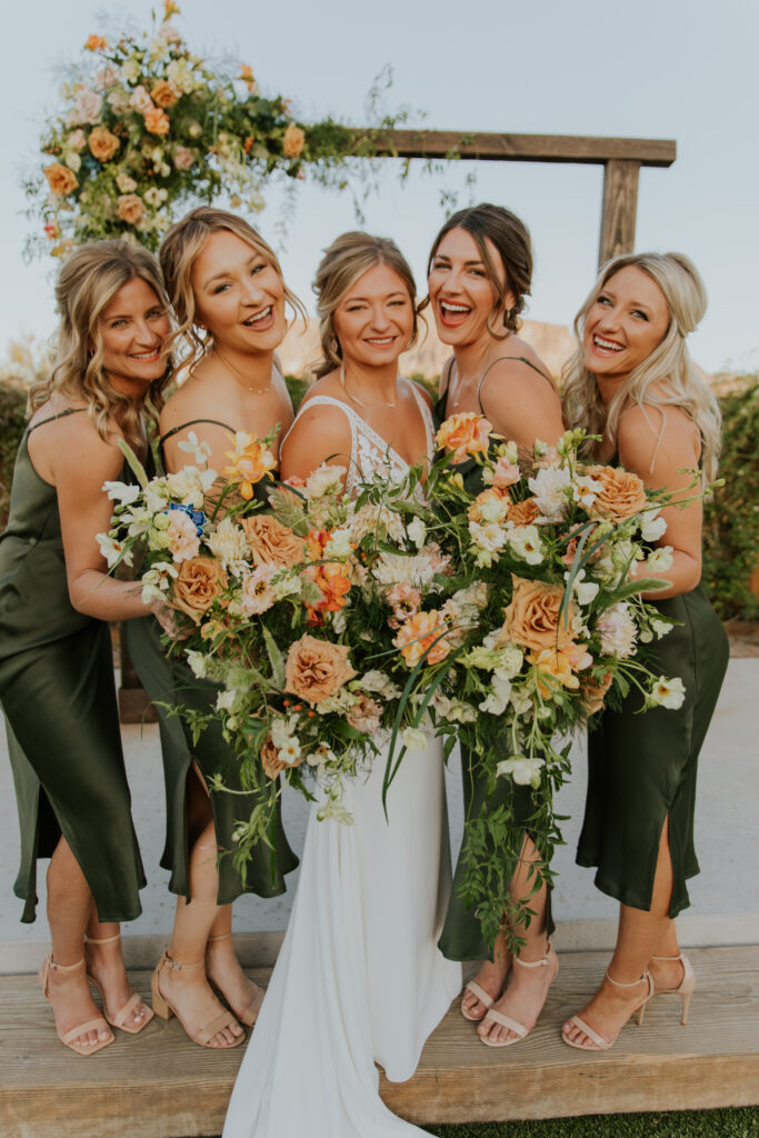 The bride and her bridesmaids smile and laugh together with their floral bouquets made of gorgeous summer flowers. #ChelseyMichelleCoPhotography #Arizonaweddingphotographer #Phoenixweddingphotographer #Apachejunctionwedding #ThePaseo #weddingatThePaseo #Gilbertphotographer #Arizonadestinationwedding