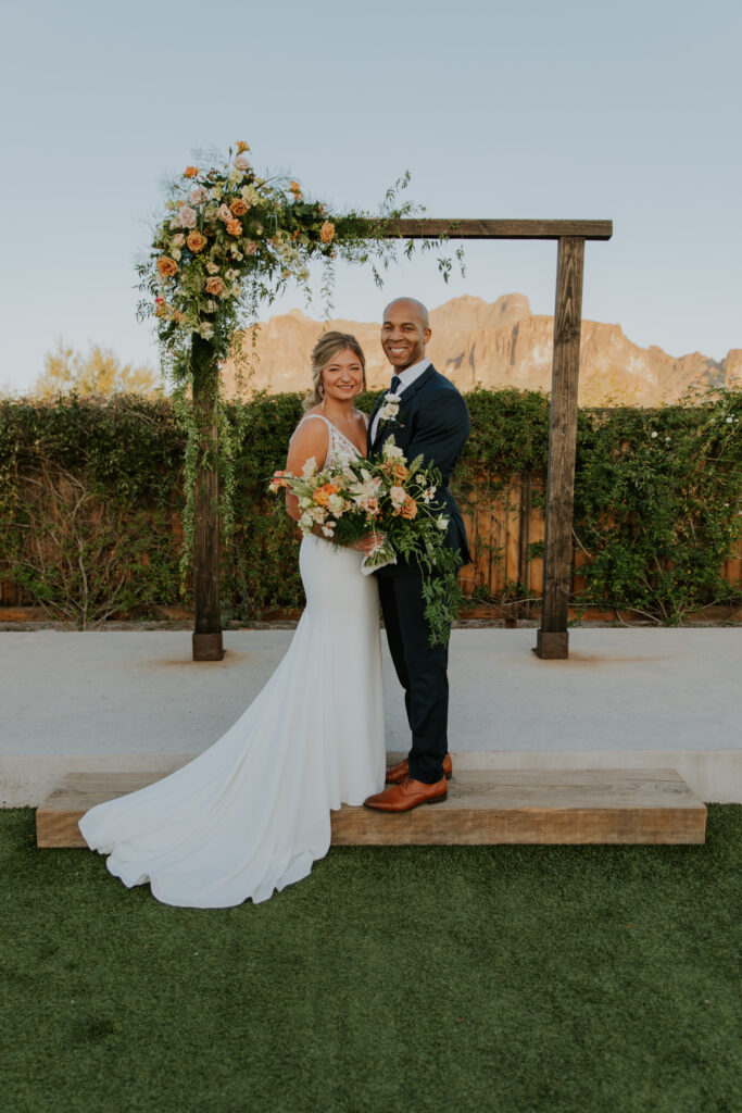 A couple shot of husband and wife standing in front of their wedding arbor decorated with a gorgeous floral arrangement. #ChelseyMichelleCoPhotography #Arizonaweddingphotographer #Phoenixweddingphotographer #Apachejunctionwedding #ThePaseo #weddingatThePaseo #Gilbertphotographer #Arizonadestinationwedding