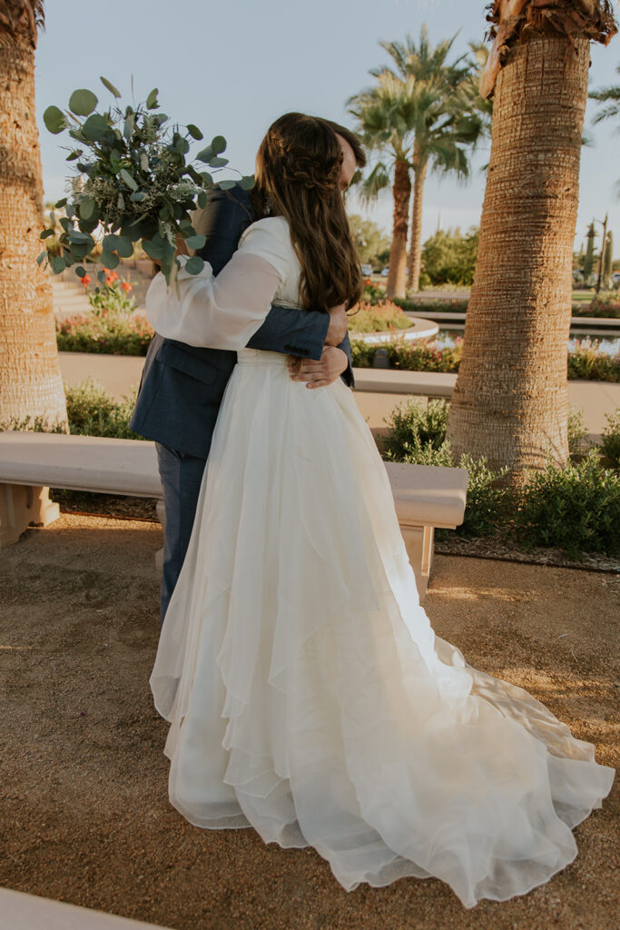 Candid first look photo at Mesa LDS temple grounds of bride and groom hugging bride wearing romantic flowing wedding dress and half up wedding hairstyle. #ChelseyMichelleCoPhotography #Arizonaweddingphotographer #Phoenixphotographer #Mesaphotographer #Gilbertphotographer #ArizonaLDSwedding