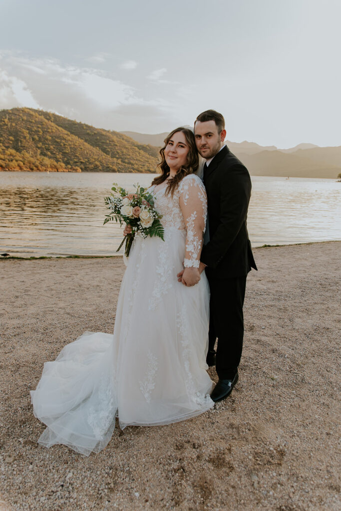 A groom tucks his bride in close and sweetly holds her hand near Saguro Lake. Bride holds a delicate, beautiful flower bouquet. 
#ChelseyMichelleCoPhotography #ArizonaWeddingPhotographer #PhoenixPhotographer #MesaPhotographer #BridalPhotos #SaguroLake
