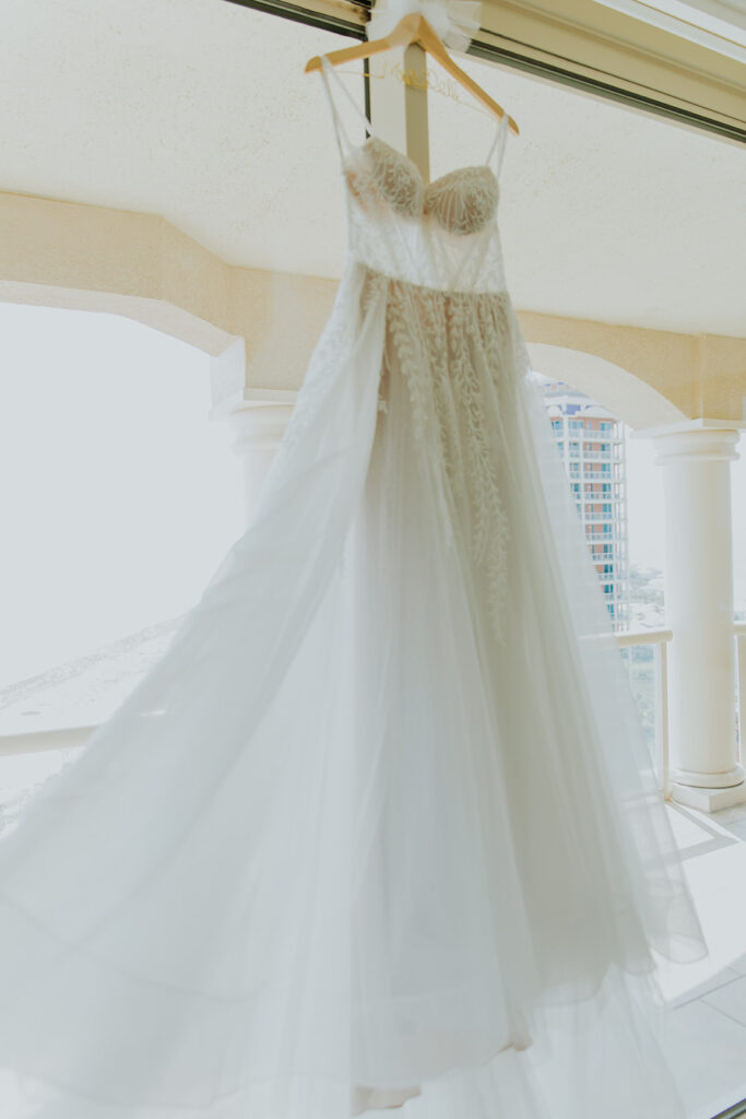 A bride’s wedding dress is beautifully displayed on a hanger in front of several large windows showcasing the delicate lace. Chelsey Michelle Co Photography travels for a destination wedding on the beach. #DestinationWedding #ChelseyMichelleCoPhotography #SummerWedding #FloridaWedding
