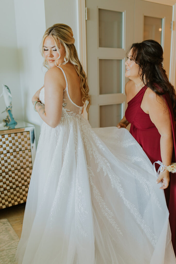 A bride stands with her mother while her mother lays out the train on her wedding dress. The bride looks over her shoulder to see all the details. Red wedding party attire. Bride prepping for wedding day. #DestinationWedding #ChelseyMichelleCoPhotography #SummerWedding
#WeddingPrep #FloridaWedding
