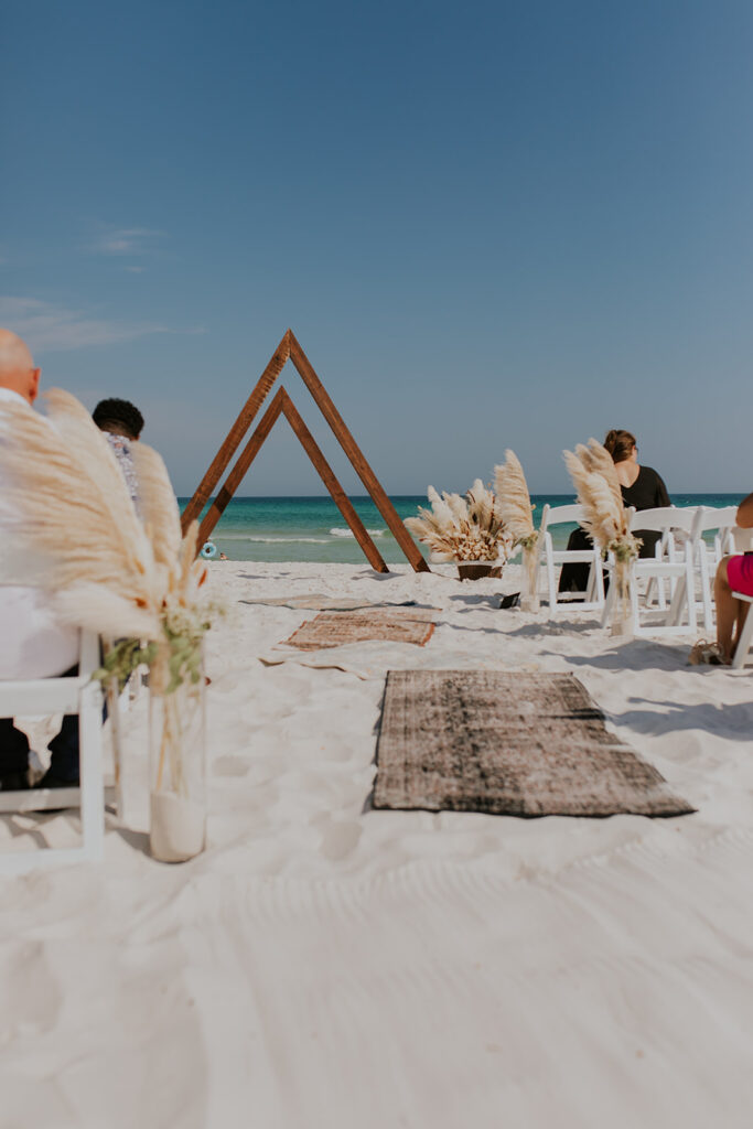 A beautiful wedding scene complete with large decorative vases. A long rug lines the aisle on the beach. There is a triangular wooden backdrop for the bride and groom to stand in front of. Chelsey Michelle Co Photography captures the details of the day. #DestinationWedding #ChelseyMichelleCoPhotography #SummerWedding #FloridaWedding
