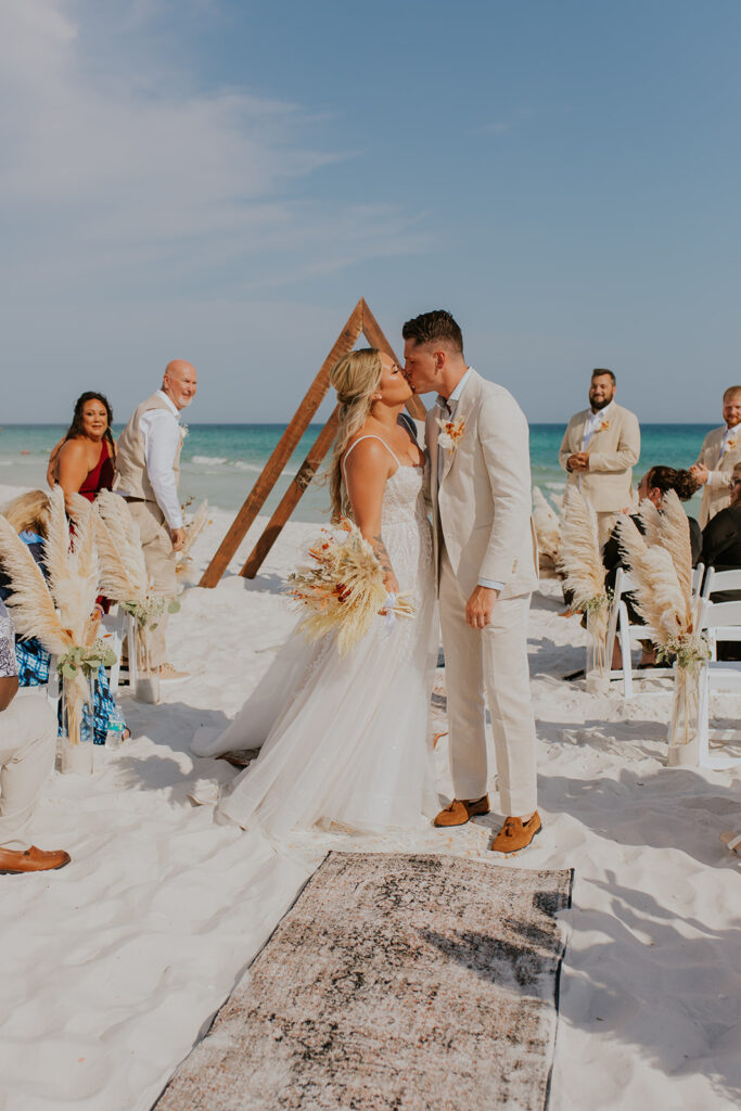 A bride and groom kiss at the end of the aisle. A beautiful wooden triangle structure is used as a backdrop. Beach wedding setup inspiration. Pensacola Beach wedding in Florida.  #DestinationWedding #ChelseyMichelleCoPhotography #SummerWedding #FloridaWedding
