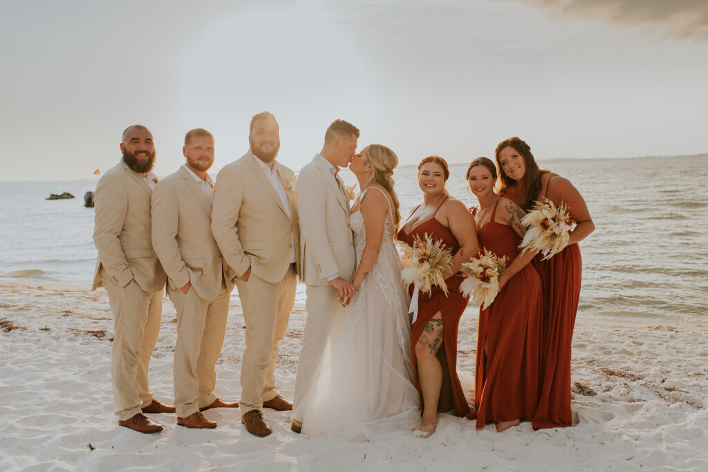 A bride and groom share a sweet kiss while surrounded by their bridal party. The groomsmen wear tan colored suits and the bridesmaids are in deep red dresses. Wedding photos on the beach.  #DestinationWedding #ChelseyMichelleCoPhotography #SummerWedding #FloridaWedding
