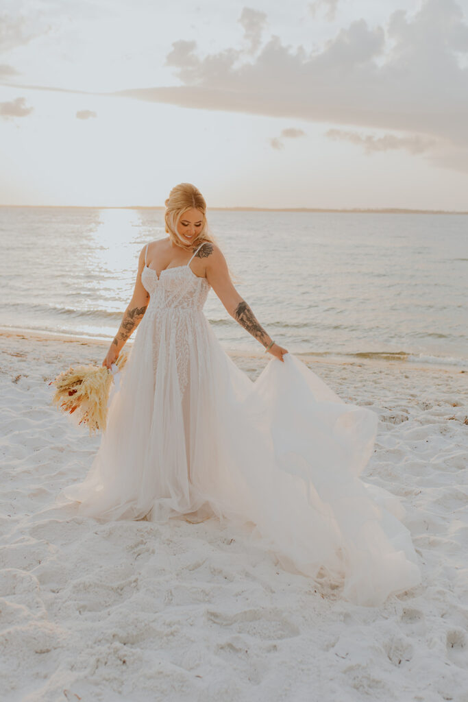 A beautiful bride stands on the beach near the ocean’s edge. She holds the train of her dress in one hand and her bouquet in the other. Chelsey Michelle Co Photography will travel for destination weddings. #DestinationWedding #ChelseyMichelleCoPhotography #SummerWedding #FloridaWedding
