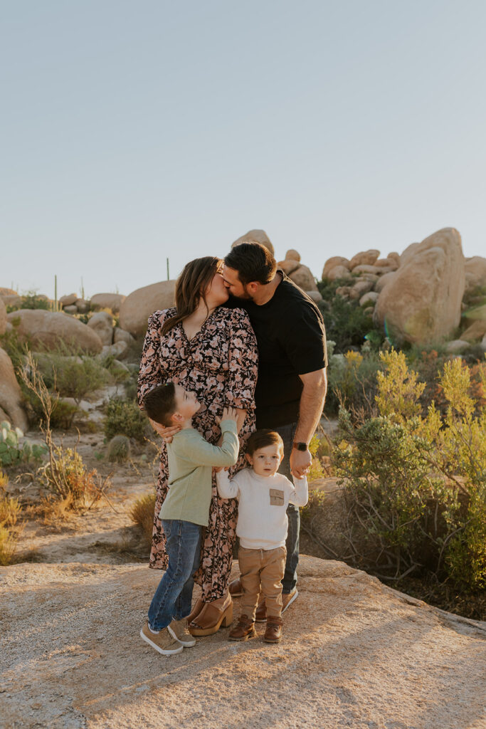 A young mom and dad share a kiss while their oldest son smiles up at them on his tip toes. Dad holds hands with the youngest son. The family stands in front of a stunning desert background.
#ChelseyMichelleCoPhotography #MaternityPhotos #EastValleyAZ #familyphotos
