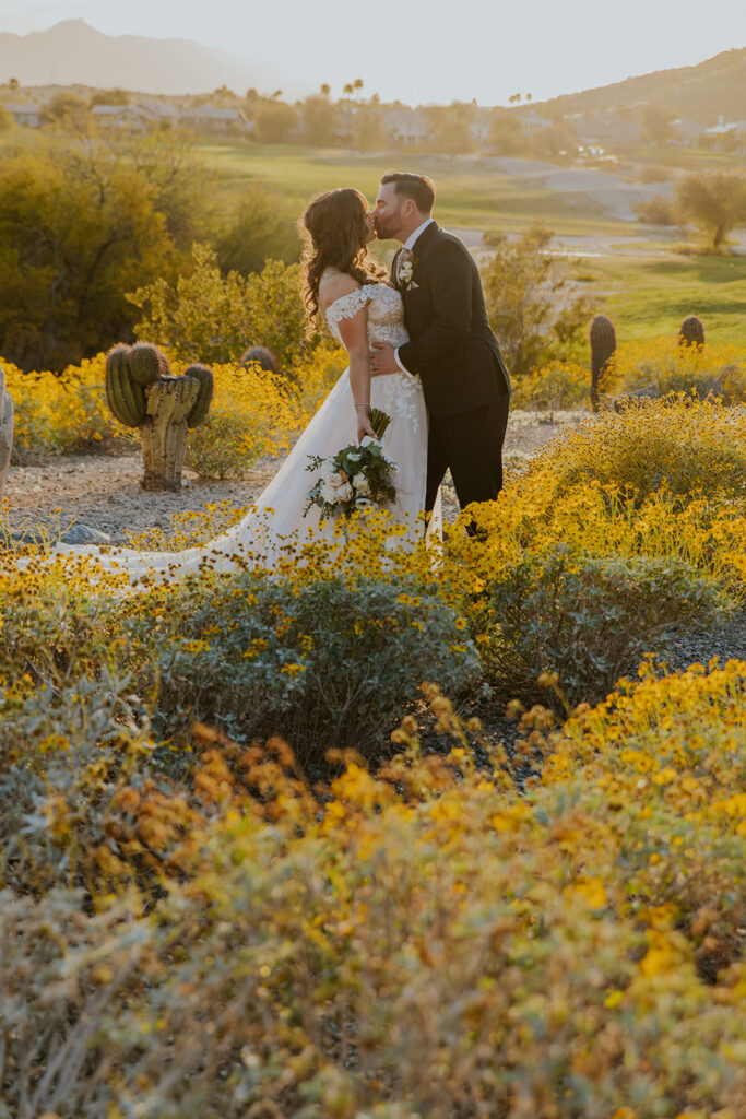 A beautifully composed photo of bride and groom kissing in the most stunning Arizona landscape, with green as far as you can see and beautiful yellow desert flowers and gorgeous desert sun. Wedding photography location landscape stunning filtered light cactus grass #Desertweddingvenues #ArizonaWeddingVenue #Arizonaweddingphotographer #Mesaweddingphotographer
