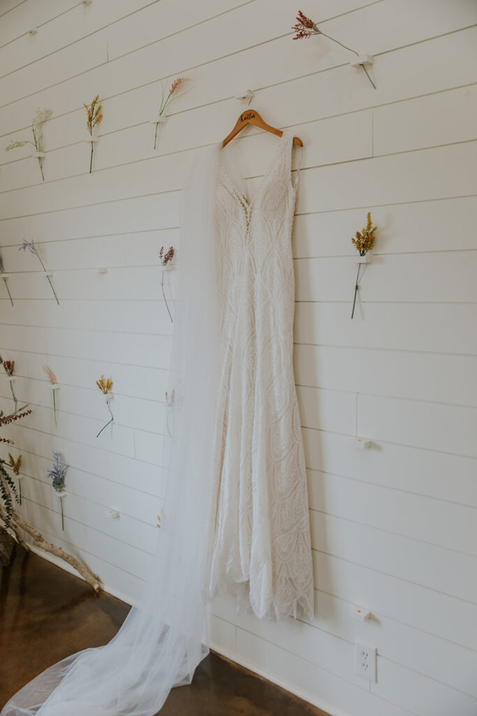 Photo showing off the most essential detail of the big day, a lacy sleeveless wedding dress hung in front of a shiplap wall with single flowers surrounding it. Wedding dress lace style fashion bride-to-be #Desertweddingvenues #ArizonaWeddingVenue #Arizonaweddingphotographer #Mesaweddingphotographer
