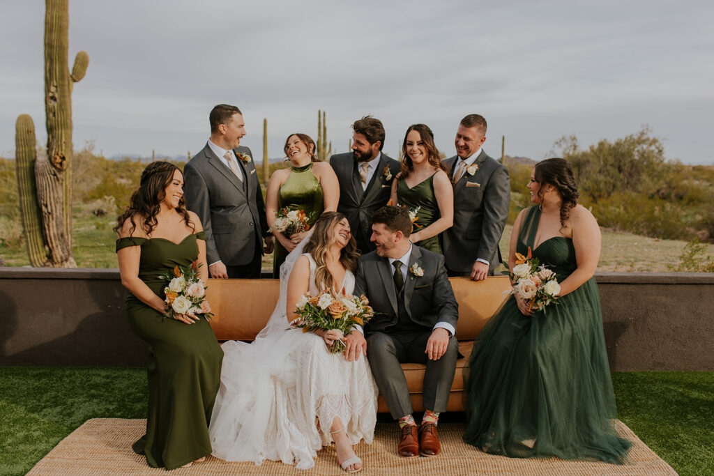 The wedding party, surrounded by desert landscape, brings together the bride's vibe through the scenery and fun leather couch and rug to stage the perfect photos. Olive hunter dark green coral florals cactus desert Arizona wedding groomsmen bridesmaids #Desertweddingvenues #ArizonaWeddingVenue #Arizonaweddingphotographer #Mesaweddingphotographer