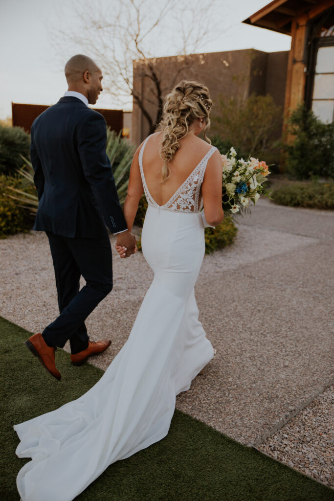 The couple walks hand-in-hand towards their venue, showcasing the stunning back of the wedding dress with stunning lace details. Scottsdale Arizona couple bride groom outfit inspiration style desert wedding #Desertweddingvenues #ArizonaWeddingVenue #Arizonaweddingphotographer #Mesaweddingphotographer
