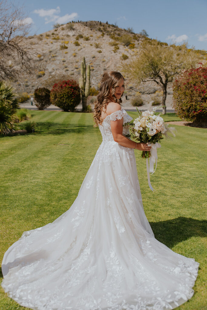 A bridal photo of rocky Arizona mountains and stunning green grass creates the perfect location to show off the stunning wedding dress. Cactus desert plants blue sky dress details fashion special day bridal bouquet inspiration #Desertweddingvenues #ArizonaWeddingVenue #Arizonaweddingphotographer #Mesaweddingphotographer