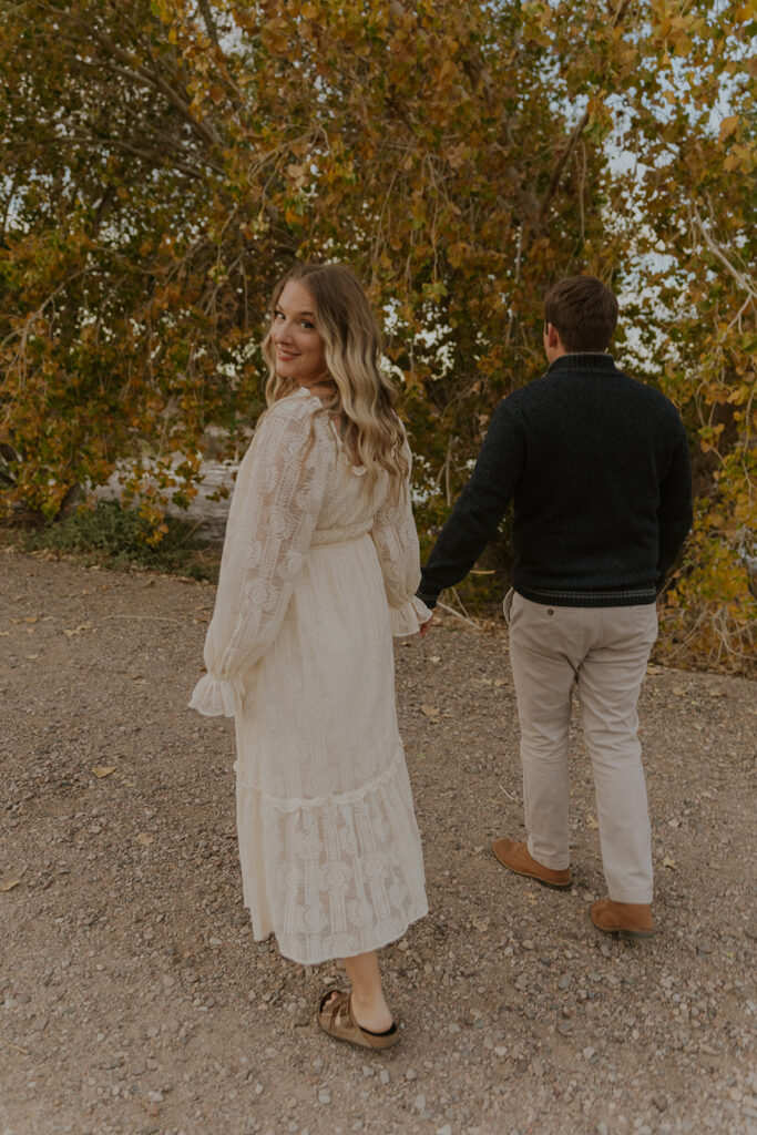 Walking together with their backs turned, a couple walks hand in hand down an Arizona dirt road while looking back and smiling. Unique engagements portraits wedding photographer #engagementphotosgilbert #azbrides #couplephotography #arizonaweddings #chelseymichellephotography
